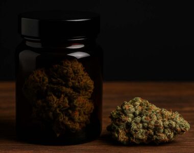 Ciemny szklany słoik z suszem marihuany na drewnianym stole, na tle czarnej ściany